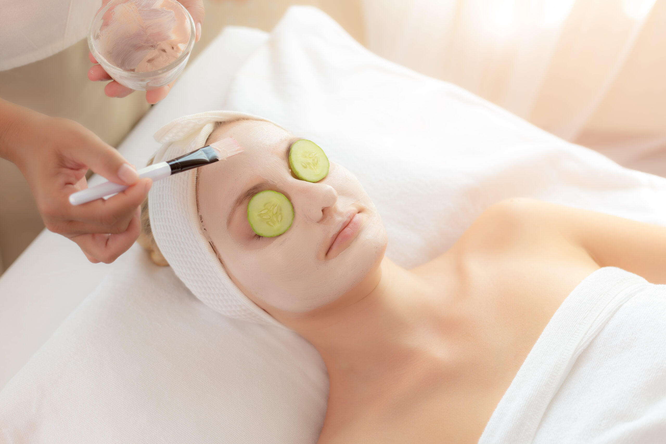 Masseuse or Massage therapist use brush for applying cream mask to beautiful customer face for healthy skin face with cucumber slices on her eyes that make hydration, refreshment and nourish skin face